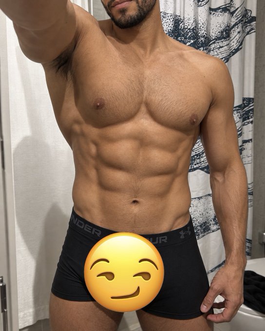 A shirtless, muscular man takes a selfie in a bathroom, wearing black Under Armour underwear with a smiley face emoji covering the front.