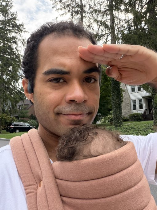 A man shields his eyes from the sun while carrying a baby in a soft pink carrier during an outdoor "GM" (good morning) selfie, embodying the sleepy, cheerful greeting vibe of the post to the @arbitrum community. The peaceful residential backdrop with trees and houses complements the casual, morning-toned crypto tweet.