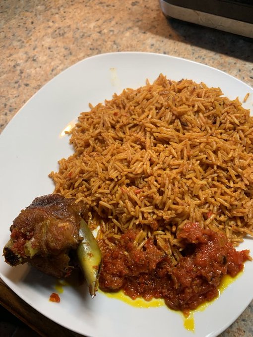 A white plate holds a serving of reddish-orange spiced rice, likely jollof or a similar dish, accompanied by a piece of meat on the bone and a chunky red tomato sauce.