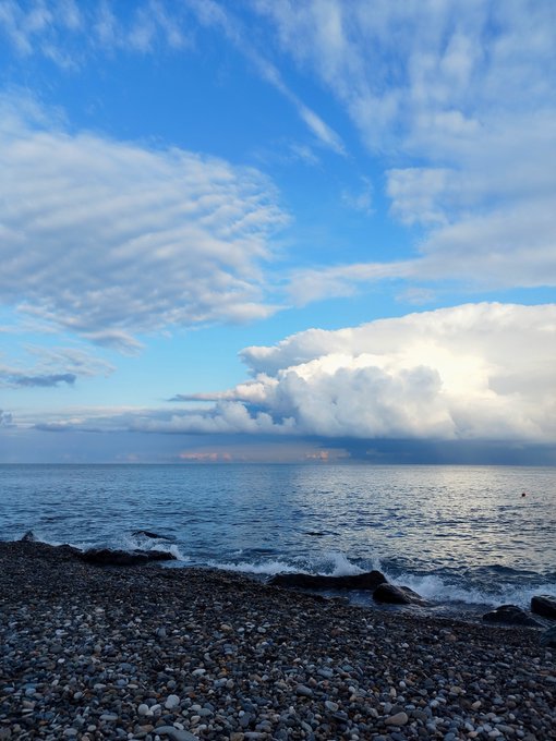 A scenic view of a rocky pebble beach meeting calm ocean waters, with dramatic white clouds filling the blue sky above.