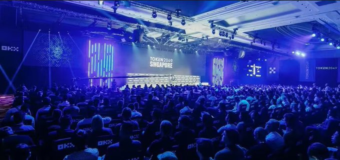 A large conference hall illuminated in blue with an audience seated facing a stage displaying 'TOKEN2049 SINGAPORE' branding.
