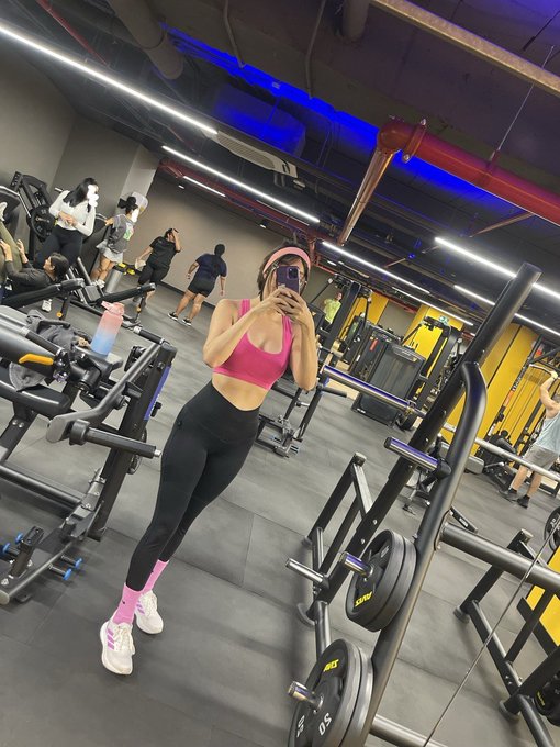 A woman in a pink sports bra, black leggings, and pink socks takes a mirror selfie in a gym with blue accent lighting, holding her phone up while resting between sets. The image captures the brief recovery moment referenced in the caption, emphasizing that rest is an essential part of any training journey.

Visible text on equipment: "STW-01" and "STW-02" labels on weight plates/equipment, with a water bottle marked with what appears to be standard gym branding.