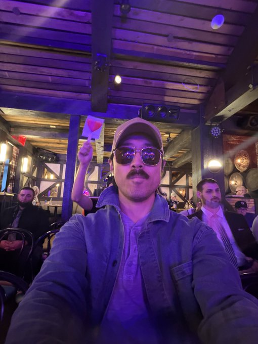 A man wearing sunglasses, a cap, and a jacket takes a selfie in a dimly lit venue with purple lighting, while a heart emoji is superimposed on the image.