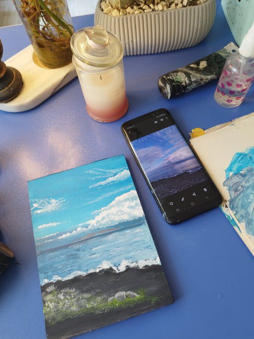 A small acrylic painting of a beach scene sits next to a smartphone displaying a reference photo of the same seascape, showcasing a self-taught artist's process of recreating an image from their screen onto canvas.