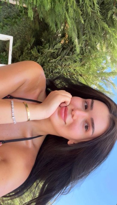 An outdoor selfie of a young woman with long dark hair, wearing a black top, silver and gold bracelets, and red lipstick, posing with her hand near her face against a backdrop of green foliage.