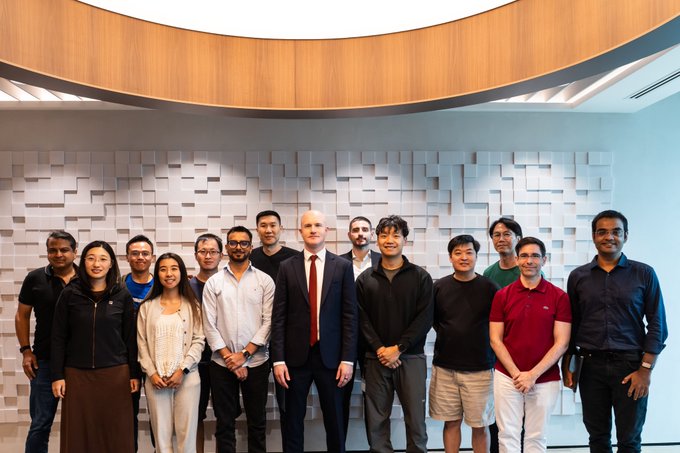 A group of approximately thirteen engineering leaders, including a man in a suit and red tie at the center, poses for a team photo inside the Coinbase Singapore office against a textured white wall. The image visually represents the post's description of a productive meeting with engineering leads focused on improving code quality and scalability.
