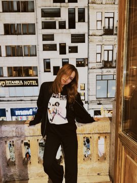 A woman wearing a black jacket and graphic t-shirt stands on a balcony with the 'TANTAWLA HOTEL' visible behind her.