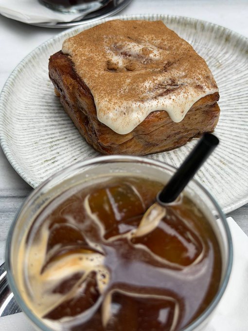 A plate with a cinnamon roll topped with cream cheese frosting sits next to a glass of iced coffee with a black straw, viewed from above.