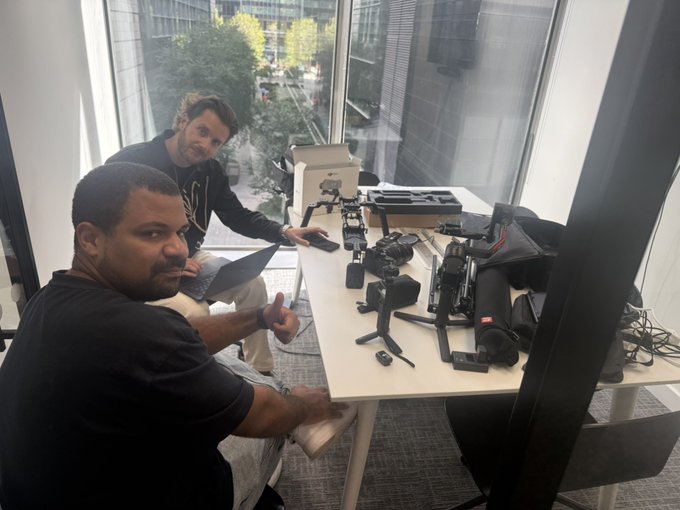 Two men sit at a desk covered with camera equipment, with one giving a thumbs-up.
