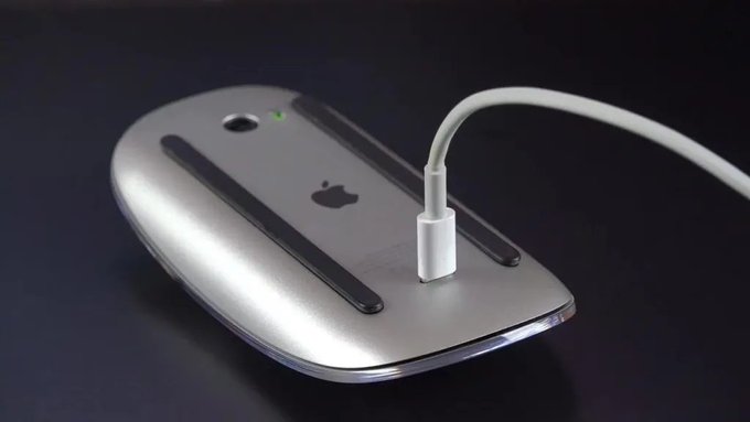 An Apple Magic Mouse is shown from below, revealing its sleek aluminum underside with the Apple logo and two black glide rails, while a white charging cable is plugged into its port.