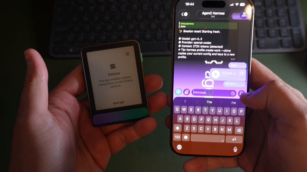 The image shows a person holding two devices - an e-reader (or tablet) and a smartphone. The person's hands are positioned over both devices, with one hand resting on each item. The scene takes place against a green background, which contrasts nicely with the devices' colors. There is also text visible in the image that reads "just told my agent(AgenC) on telegram: send 0.1 SOL from my ledger to this address" and "focus on what is in the image and how it relates to the post".