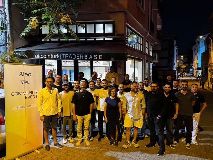 A group of people pose for a photo outside a venue labeled 'altcointurk | TRADER BASE' at night, with a banner next to them reading 'Aleo Community Event Istanbul, Turkey'.