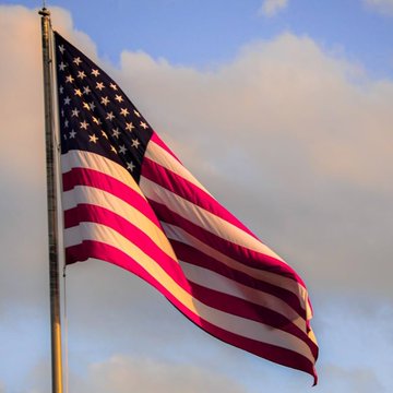 An American flag waving on a flagpole against a blue sky with scattered clouds, illuminated by warm golden light.