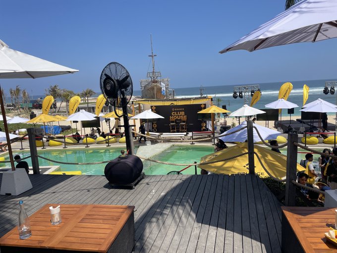 A beachside club venue called 'CLUB HOUSE' with a pool, yacht-shaped stage, large fan, white umbrellas, and yellow accents, viewed from a wooden deck with tables in the foreground.