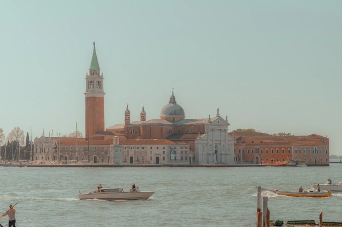 A view of Venice featuring the iconic San Giorgio Maggiore church and its tall bell tower across the water, with small boats navigating the lagoon.
