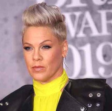 A woman with a short platinum blonde pixie cut and a yellow high-neck top poses at an event with "WAF" and "019" visible in the background. The image appears to show her at a 2019 awards ceremony, likely shared in a post celebrating reaching 111K signatures.