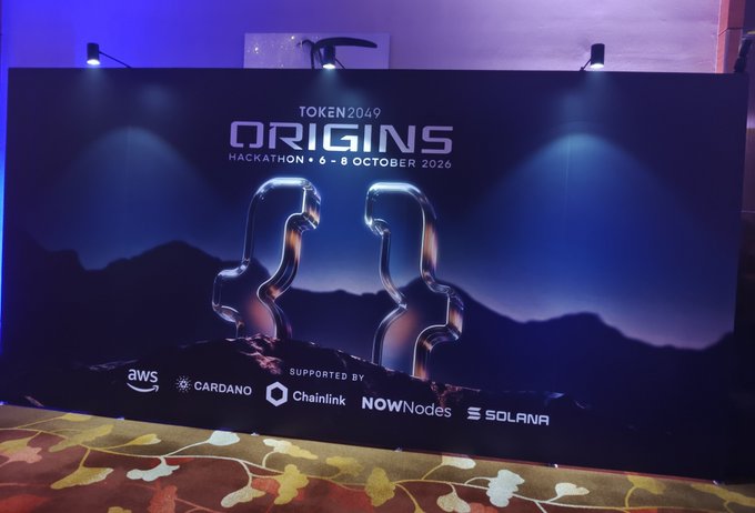 A promotional banner for 'TOKEN2049 ORIGINS HACKATHON • 6-8 OCTOBER 2026,' supported by AWS, Cardano, Chainlink, NOWNodes, and Solana, featuring an artistic design of two glass-like figures facing each other.
