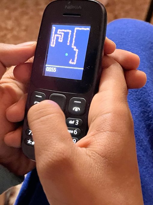 A close-up photo of hands holding a classic black Nokia phone playing the game Snake, with the score '0155' visible on the small blue screen.
