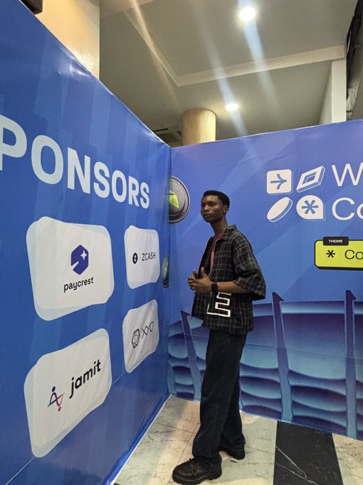 A man in a plaid shirt stands at a tech event in front of blue sponsor walls displaying logos for 'paycrest', 'Zcash', and 'jamit' under the heading 'SPONSORS'.