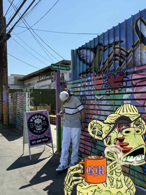 A man in a white shirt and cap looks at his phone next to a colorful graffiti-covered shipping container, with a "EVEN FLOW BREWING - WE ARE OPEN" sign displayed.