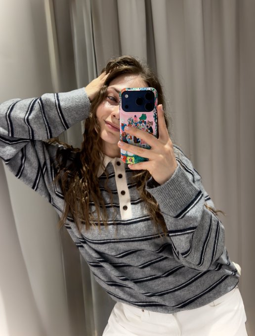 A young woman with long wavy hair takes a mirror selfie wearing a gray striped long-sleeve polo and white pants, holding a phone with a colorful illustrated case in what appears to be a fitting room with curtains behind her. The image reflects the personal, relatable vibe of her post about being a 26-year-old content creator in the web3/crypto space, showcasing her approachable, creative personality as she seeks to connect with founders and builders in the industry.
