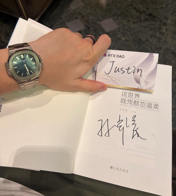 A hand wearing a watch holds a name card reading 'HTX DAO Justin' above a signed Chinese book titled '这世界既残酷也温柔'.