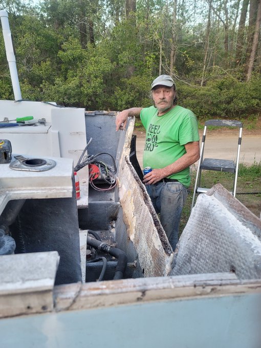 A man in a green t-shirt and cap standing next to the exposed engine compartment of a boat.