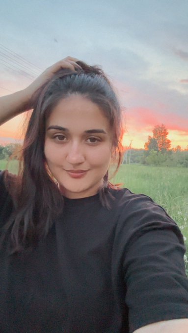 A young woman smiles for a selfie in a green field during a vibrant sunset, wearing a black long-sleeved shirt with her hair pulled back.