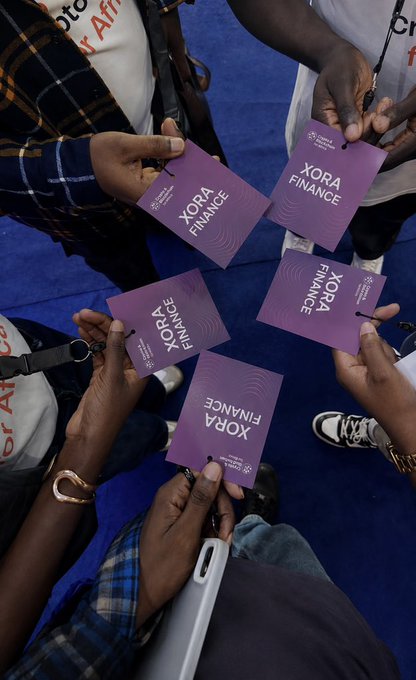 An overhead shot of multiple hands holding purple cards and lanyards labeled 'XORA FINANCE' and 'Africa Blockchain Center' at what appears to be a conference or event with blue carpeting.