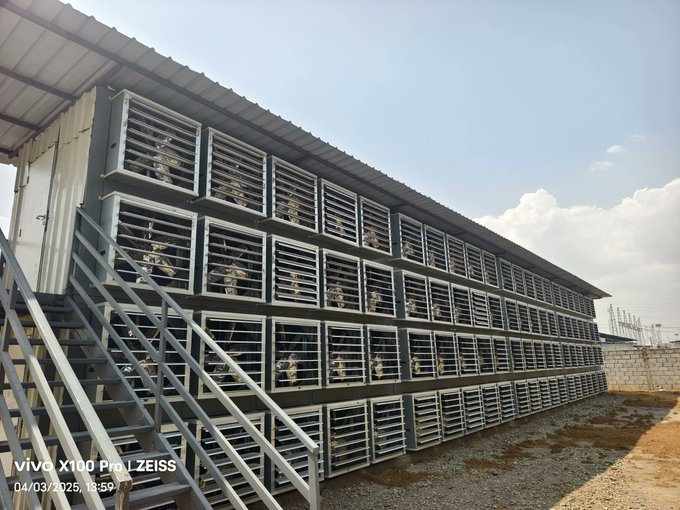 An outdoor industrial setup of stacked metal cages, likely for cooling or equipment housing, with stairs on the left side, photographed with a vivo X100 Pro on 04/03/2025.
