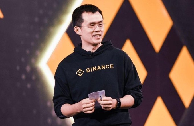 A man wearing glasses and a black "BINANCE" hoodie stands on stage holding a card, appearing to speak at an event. The image relates to the post by showing CZ, the founder of Binance, whose early Bitcoin investment helped build the cryptocurrency empire he now leads.