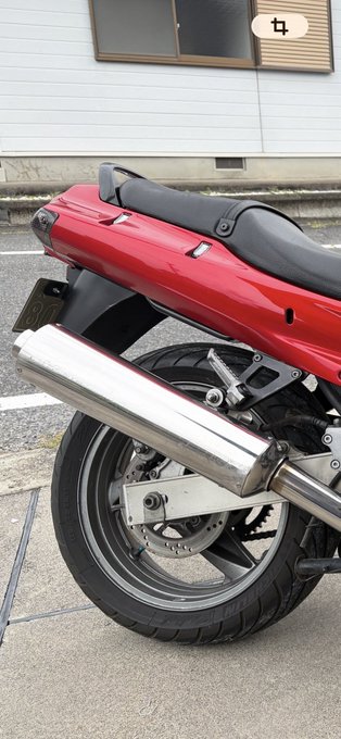 A close-up of the rear section of a red motorcycle parked on a street, featuring a black seat, silver exhaust pipe, and rear wheel with disc brake.