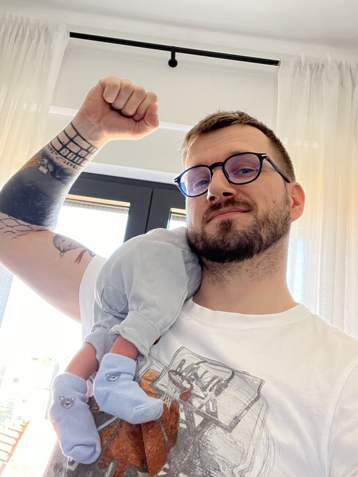 A bearded man wearing glasses and a graphic t-shirt poses with a raised, flexed fist while holding a small, blue-clothed baby doll under his other arm.