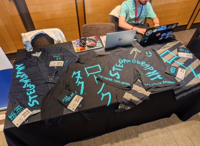 A merchandise table at a convention displaying black denim shirts and t-shirts with teal graffiti-style graphics and Japanese characters, alongside a HP laptop and stickers.