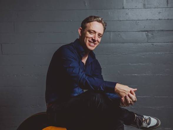 A man with dark hair wearing a dark blue shirt and black pants sits smiling on a stool against a dark, textured background.