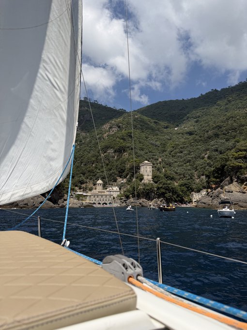 A serene morning view from a sailboat approaching a coastal village nestled against lush green hills, with historic buildings and a tower visible along the shoreline, perfectly capturing a peaceful "Gm" at sea.