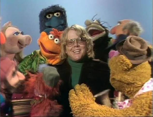 A scene from the Muppets shows a man with blonde hair and glasses smiling while surrounded by several puppet characters, including Miss Piggy, Ernie, and Fozzie Bear.