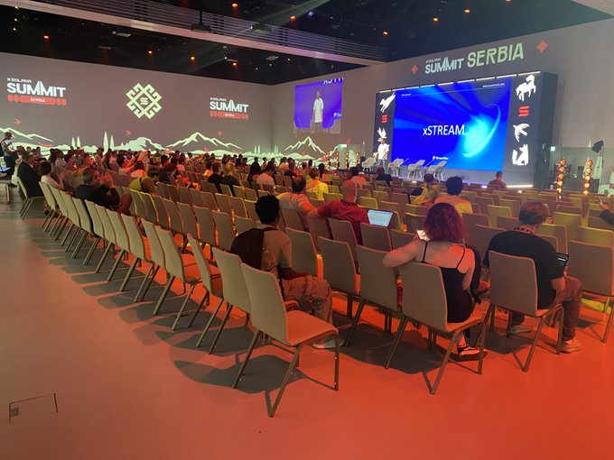 Photograph of the XLayer Summit Serbia 2026 conference, showing a large seated audience facing a stage with bright screens displaying 'xSTREAM' content.