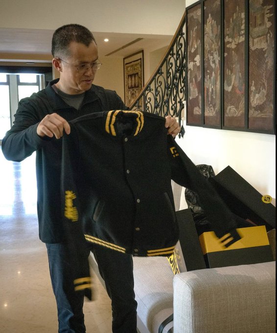 A man wearing glasses displays a black varsity jacket with yellow trim and patches, holding it up in an ornately decorated room.