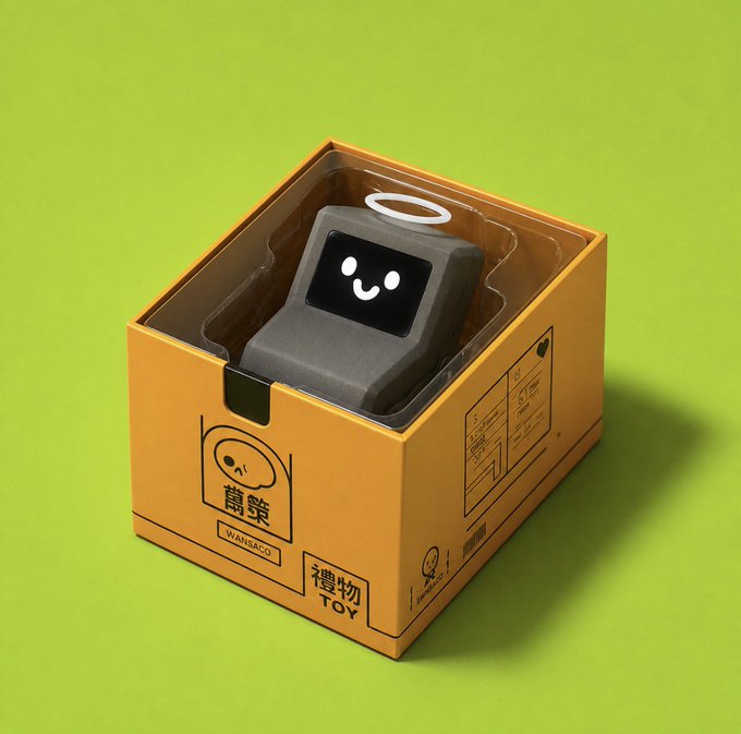 A small gray toy robot with a smiling face and a halo sits inside a yellow cardboard box with Japanese text and the word 'TOY' printed on it.