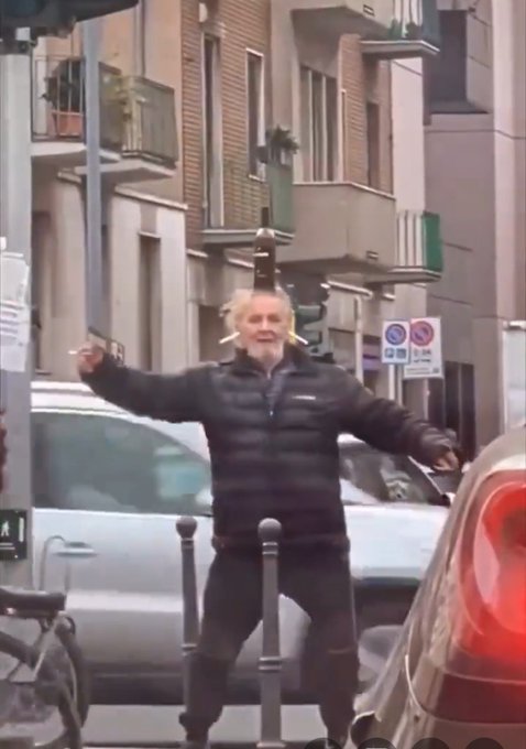 An older man with white hair stands on a city street with his arms outstretched, smiling broadly as parked cars and buildings appear in the background.