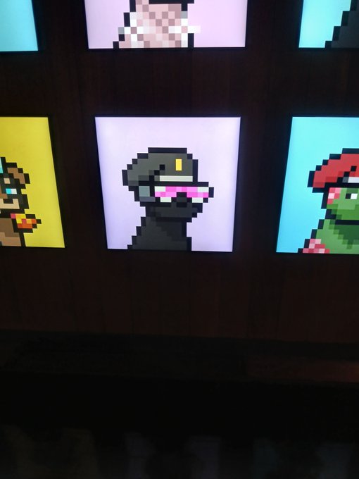 Display of pixel art portraits on illuminated panels, featuring a central character with pink sunglasses on a pink background, flanked by other characters on yellow and blue backgrounds.