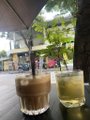 Two drinks on a glass table at a café or restaurant, one appearing to be an iced coffee and the other a yellow beverage, with a street view visible through the window.