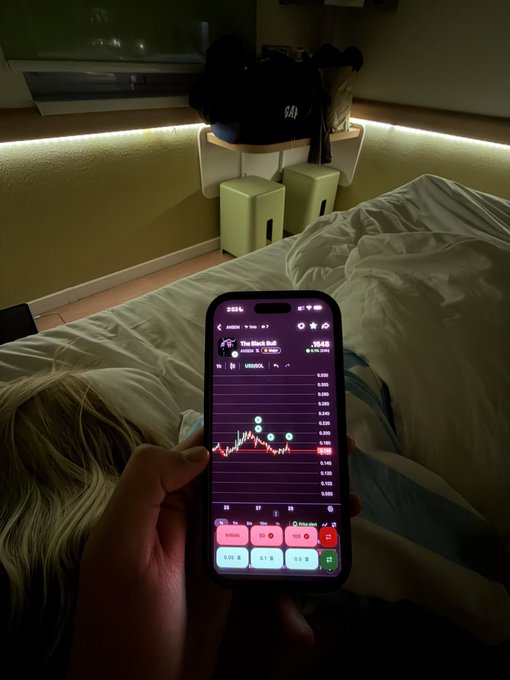 A phone displays a "BLACK KILL" stock chart showing a downward-trending graph in a dimly lit bedroom, illustrating the $ANSEM critique that it is "due for a major reversal soon."
