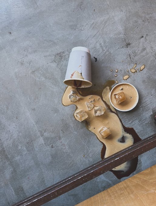 A spilled coffee cup lies on a gray concrete surface, with the brown liquid and ice cubes spread across the floor alongside a small ceramic cup.