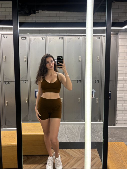 A woman takes a mirror selfie in a gym locker room, wearing a brown sports bra and matching high-waisted shorts, with lockers numbered 118, 119, 120, 151, 152, 84, and 88 visible behind her, ready to "smash the day" with her workout.