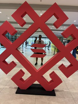 A large red geometric sculpture resembling an interlocking cross pattern stands indoors in what appears to be a shopping mall or exhibition space.