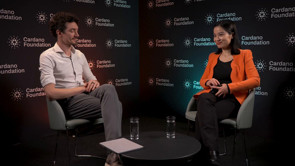 The image shows a man and woman sitting across from each other at a table with water glasses placed between them, appearing to be engaged in conversation. The background features a black wall adorned with various logos, including "Cardano Foundation" and "Metaverse Institute".