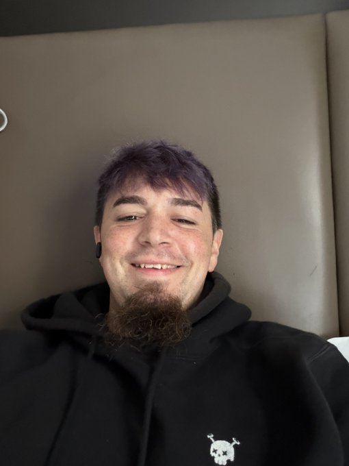 A smiling man with purple hair and a beard, wearing a black hoodie with a small skull logo, reclining against a tan cushioned surface.