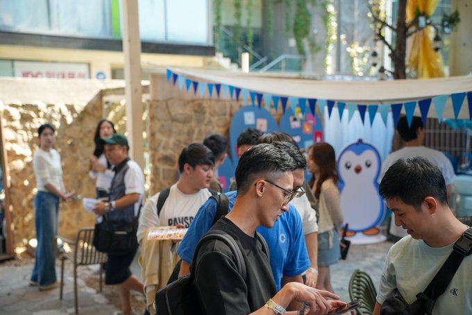 The image shows a group of young people gathered at what appears to be an outdoor pop-up event or community space, with a "PAUSE" t-shirt visible and a penguin mascot decoration in the background. A man in the foreground is focused on his phone while others mingle around decorative bunting and a yellow banner, reflecting the social, community-focused nature of "Korea Huddle."
